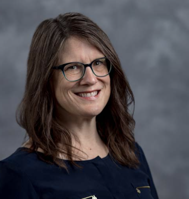 A person with long brown hair and glasses, wearing a navy blue top, smiles at the camera. The background is a blurred, neutral gray color.